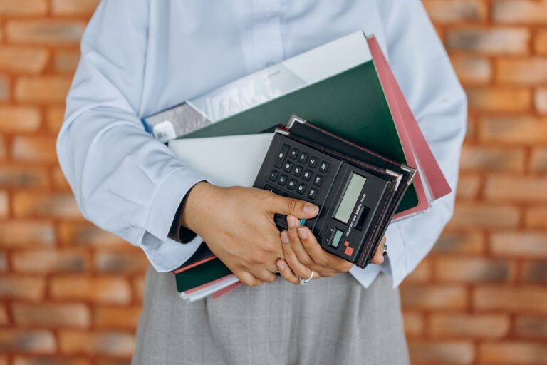 bookkeeping services small business — close-up of a person holding a calculator and folders against a brick wall background.